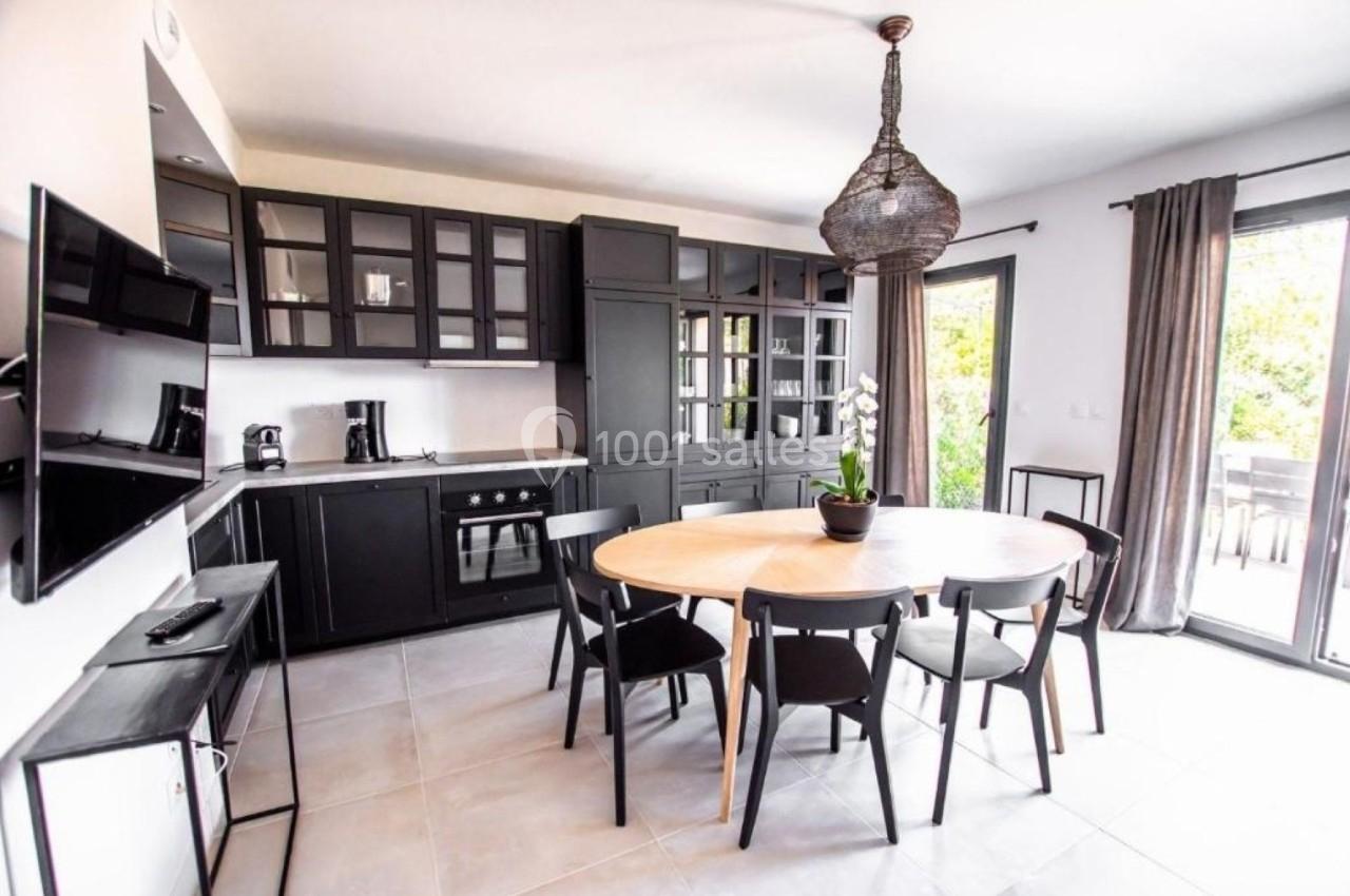 Cuisine moderne avec meubles noirs, table ronde en bois entourée de chaises, et grande fenêtre donnant sur l'extérieur.