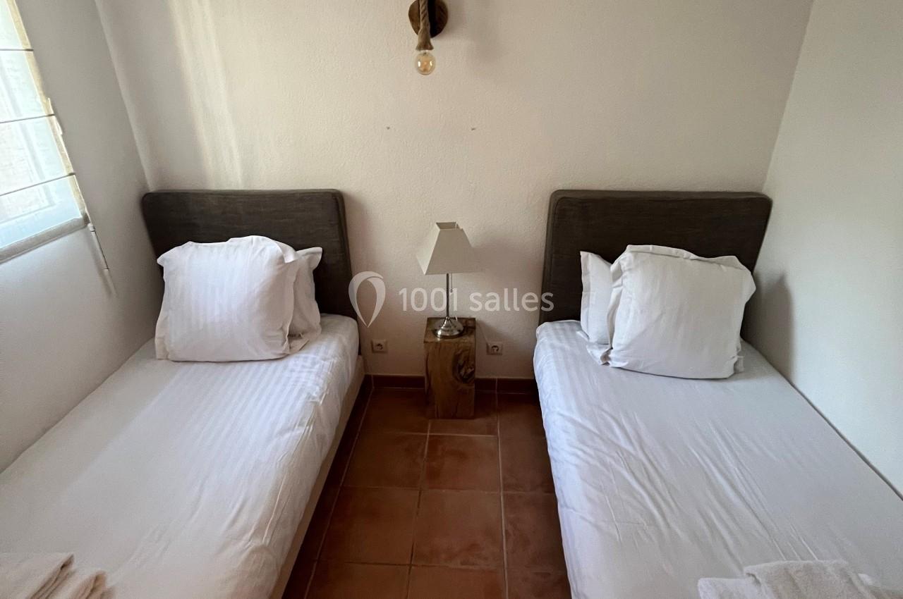 Deux lits simples avec draps blancs, une lampe de chevet centrale et un sol en carrelage dans une chambre lumineuse.