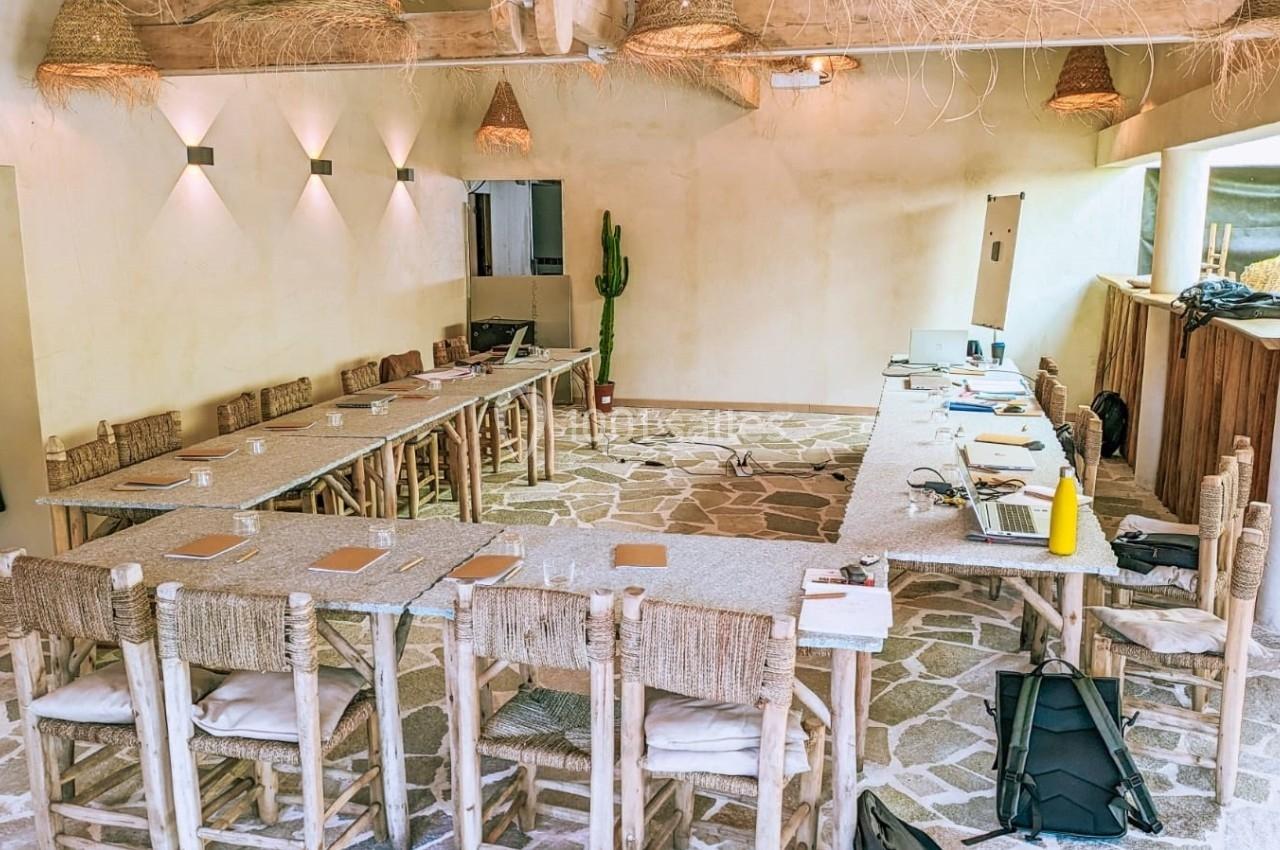 Salle de réunion lumineuse avec tables en U, chaises en bois et décoration naturelle, sol en pierre et cactus en arrière…