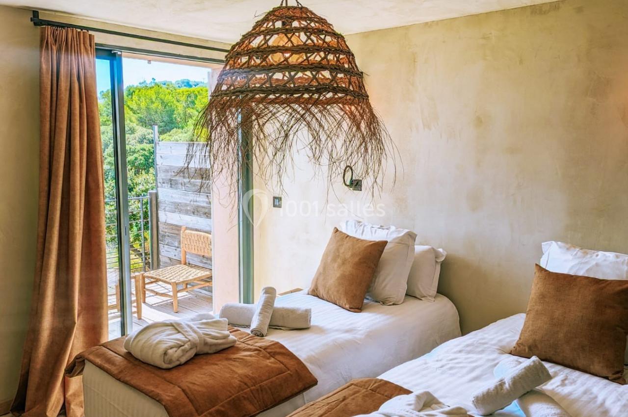 Deux lits simples avec coussins bruns dans une chambre lumineuse, ouverte sur une terrasse avec vue sur la végétation.