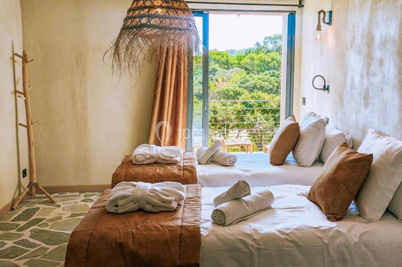 Chambre lumineuse avec deux lits simples, linge de maison beige, grande fenêtre donnant sur un paysage verdoyant.