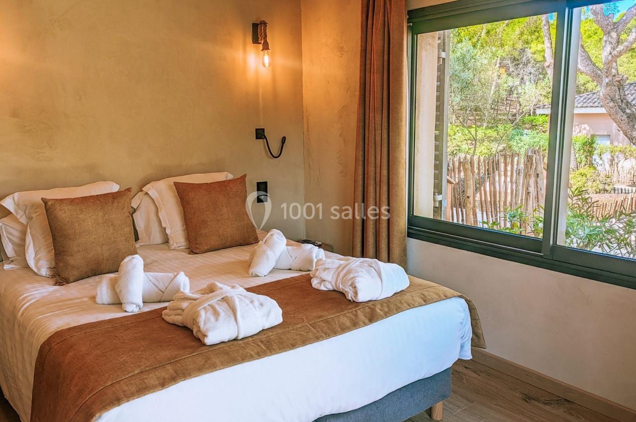 Chambre lumineuse avec lit double, oreillers et peignoirs, fenêtre donnant sur un jardin arboré.