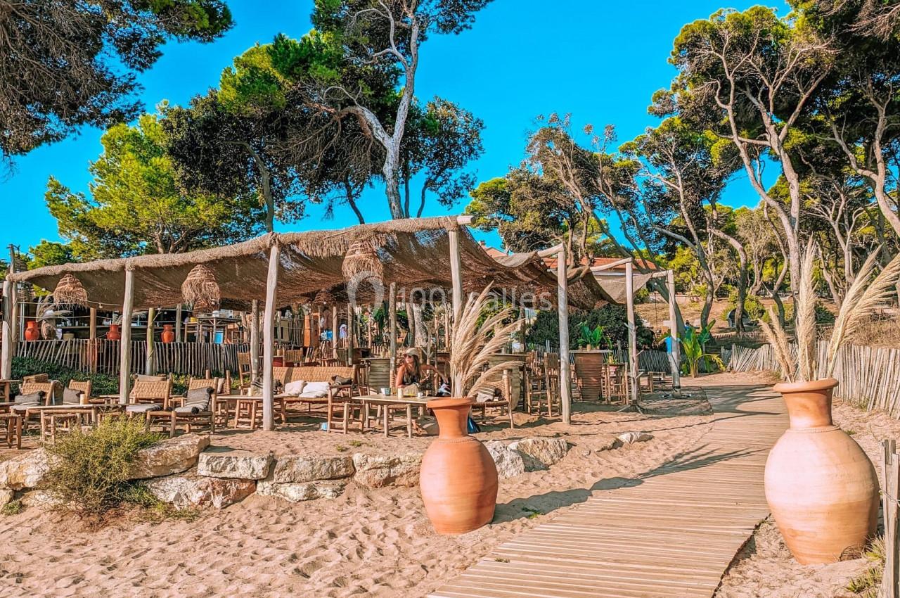 Terrasse en bois ombragée avec des tables et chaises, entourée de pins, accessible par un chemin sablonneux.