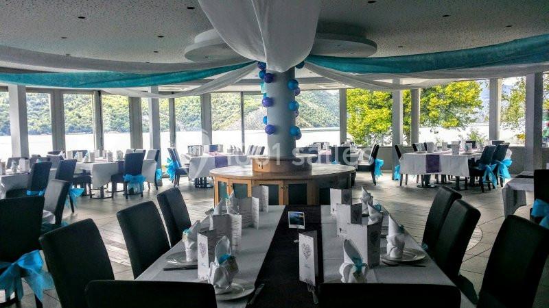 Salle de réception lumineuse avec tables dressées, nappes blanches et décorations bleues, offrant une vue sur un lac.