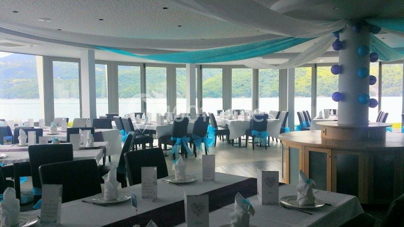 Salle de réception lumineuse avec tables dressées, décorations bleues et vue panoramique sur un lac et des collines.