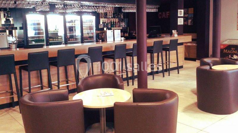 Bar avec comptoir en bois, tabourets hauts, tables rondes et fauteuils en cuir dans un espace lumineux.