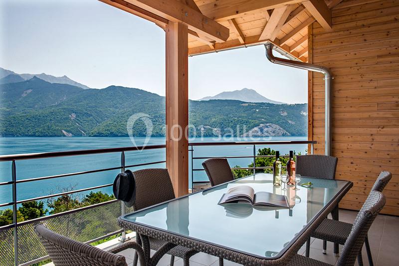 Terrasse en bois avec table en verre, chaises en rotin, vue sur un lac entouré de montagnes et ciel dégagé.
