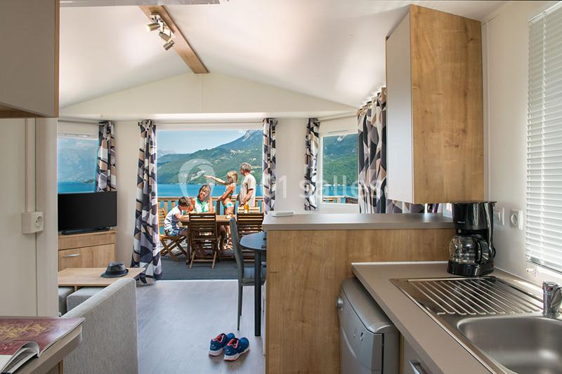 Intérieur d'un mobil-home avec cuisine équipée, vue sur une terrasse où des personnes profitent d'un paysage montagneux.