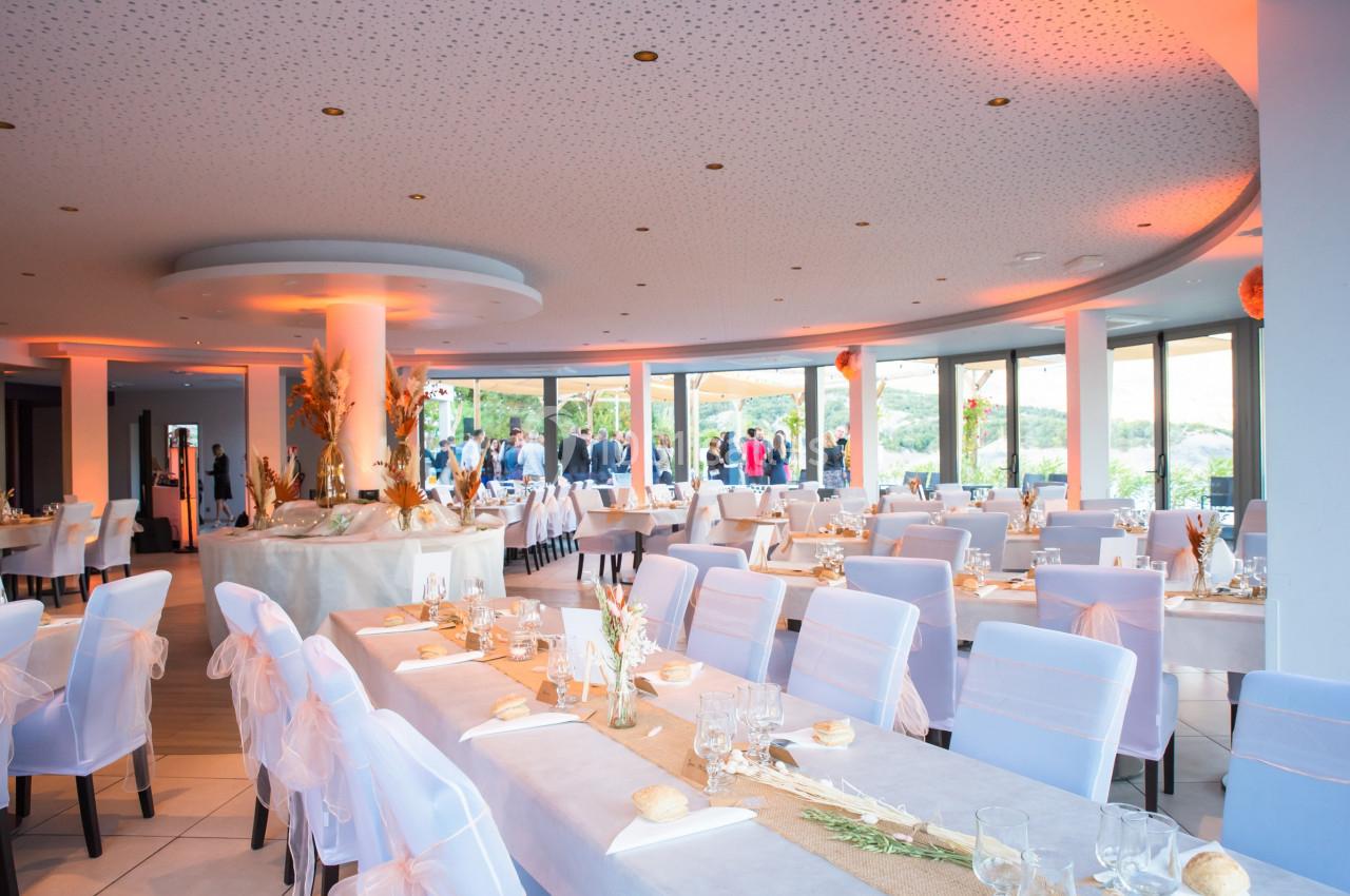 Salle de réception lumineuse avec tables dressées, chaises décorées et grandes baies vitrées donnant sur l'extérieur.