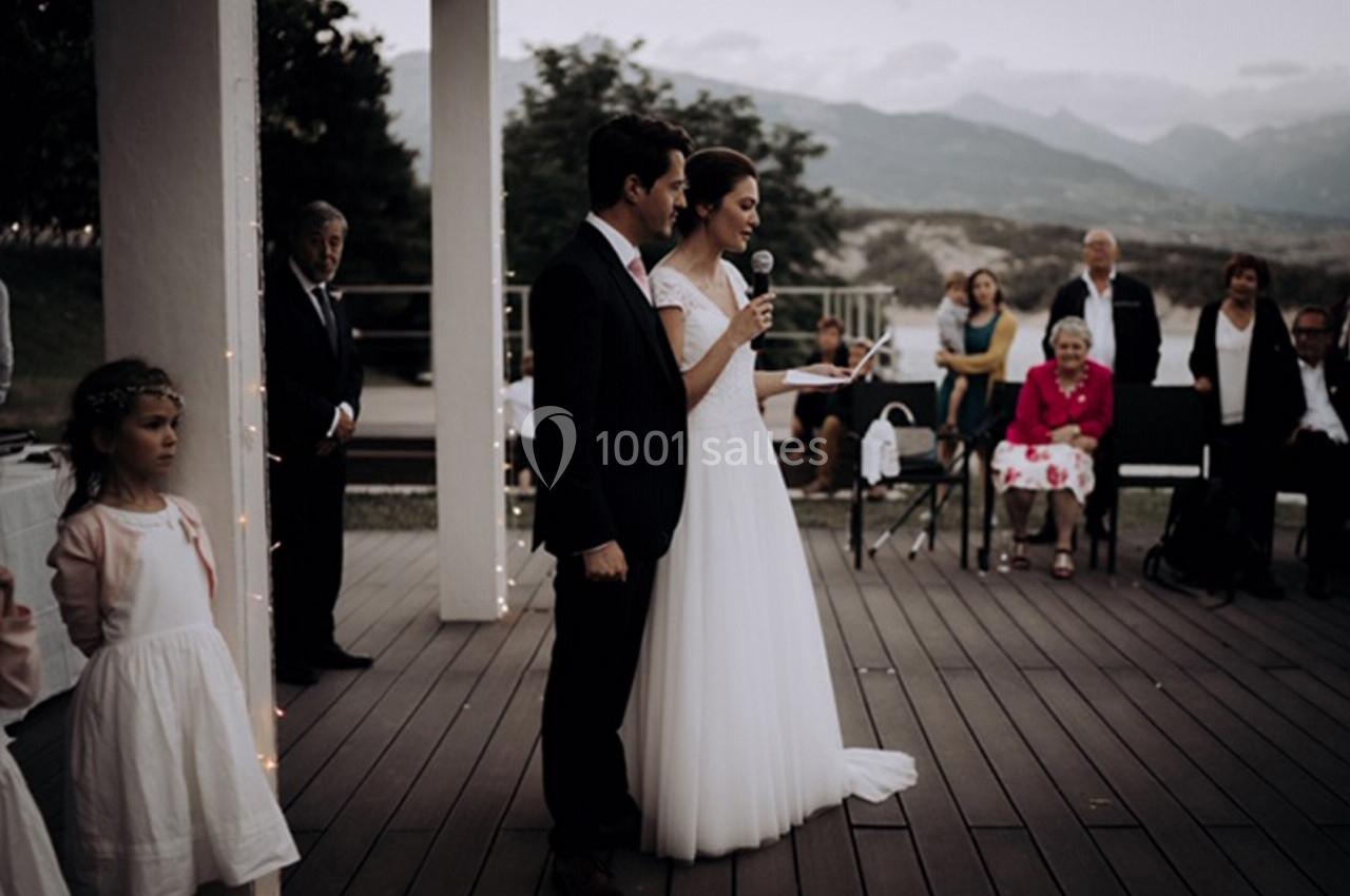 Un couple en tenue de mariage prononce un discours sur une terrasse en présence d'invités assis et debout.