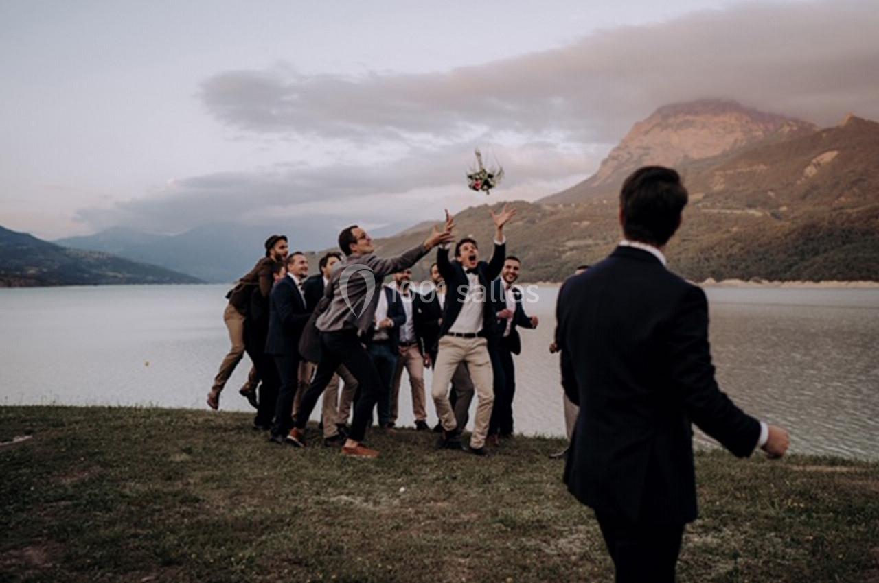 Des hommes en costume tentent d'attraper un bouquet lancé près d'un lac, avec des montagnes en arrière-plan.