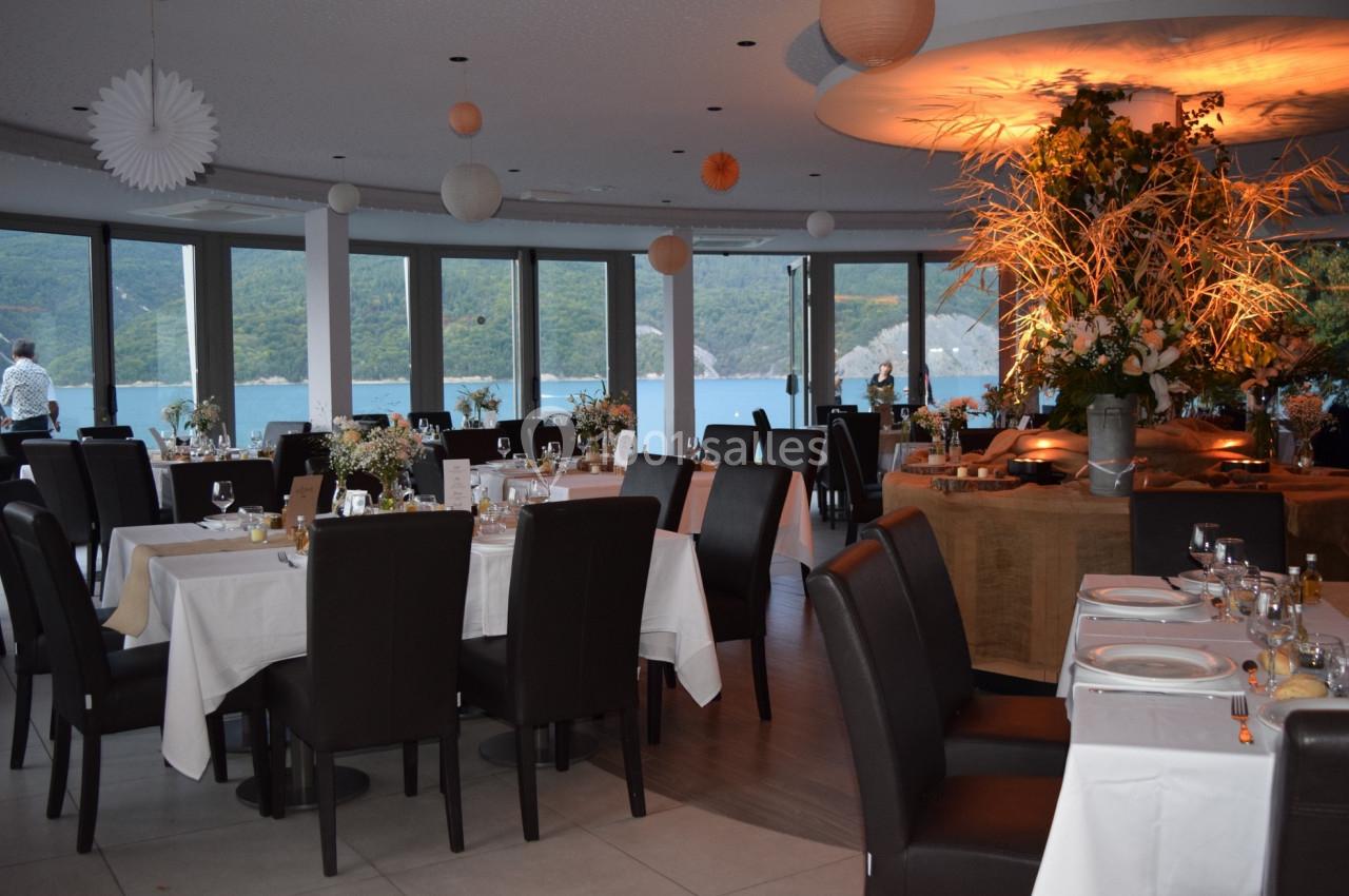 Salle de restaurant élégante avec grandes baies vitrées donnant sur un paysage de lac et montagnes, tables dressées.