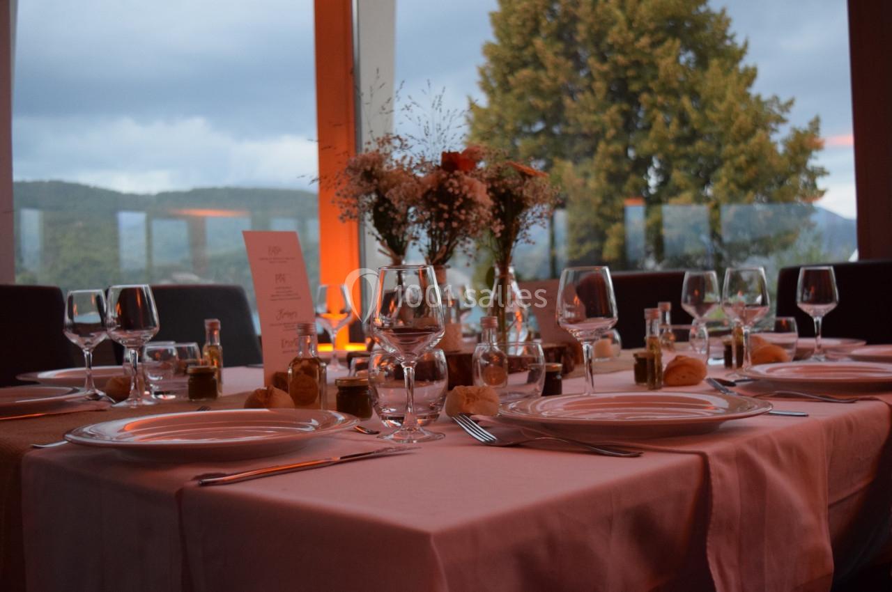 Table dressée avec nappes roses, vaisselle élégante, fleurs au centre et vue sur un paysage boisé à travers de grandes…