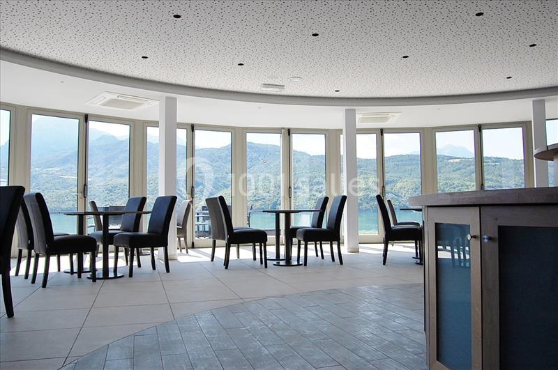 Salle lumineuse avec grandes baies vitrées offrant une vue sur un paysage montagneux et un lac, tables et chaises disposées.