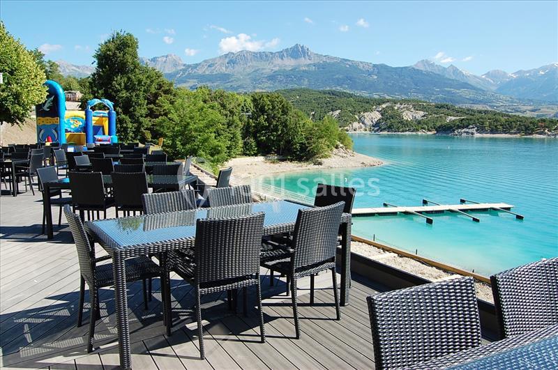 Terrasse avec tables en rotin noir surplombant un lac turquoise, entourée de montagnes et de verdure.