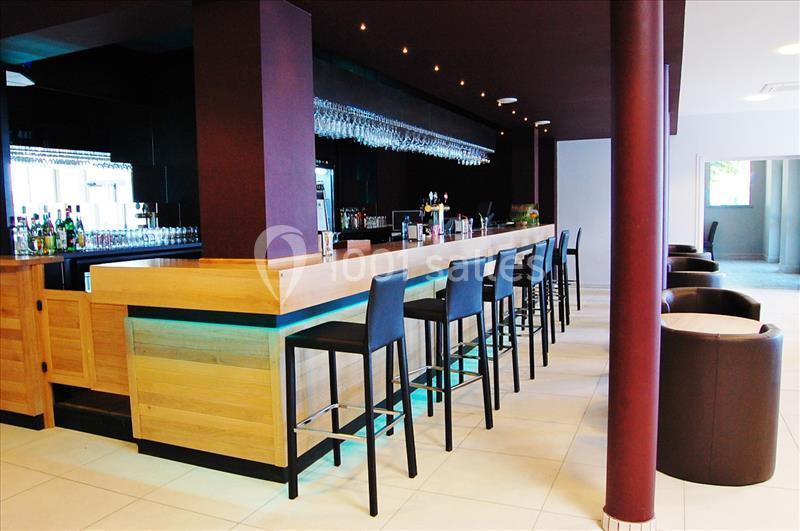 Bar moderne avec comptoir en bois clair, tabourets noirs et rangée de verres suspendus dans un espace lumineux.