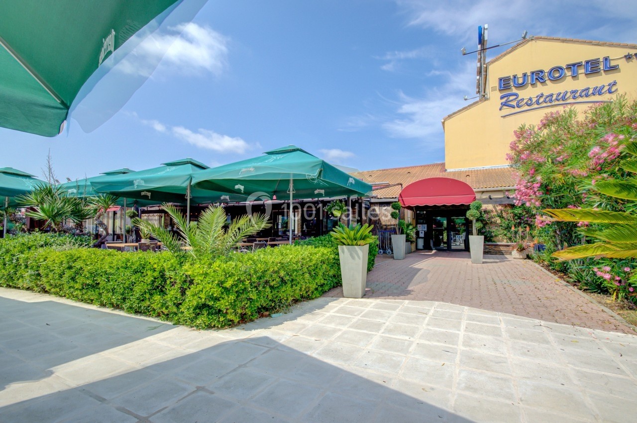 Entrée d'un restaurant avec terrasse ombragée par des parasols verts, entourée de végétation et d'un ciel dégagé.