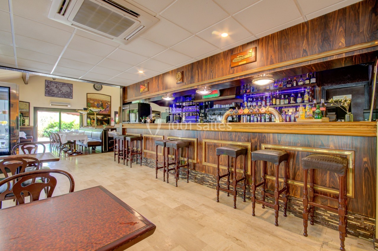 Intérieur d'un bar avec comptoir en bois, tabourets alignés, étagères de bouteilles et lumière tamisée.
