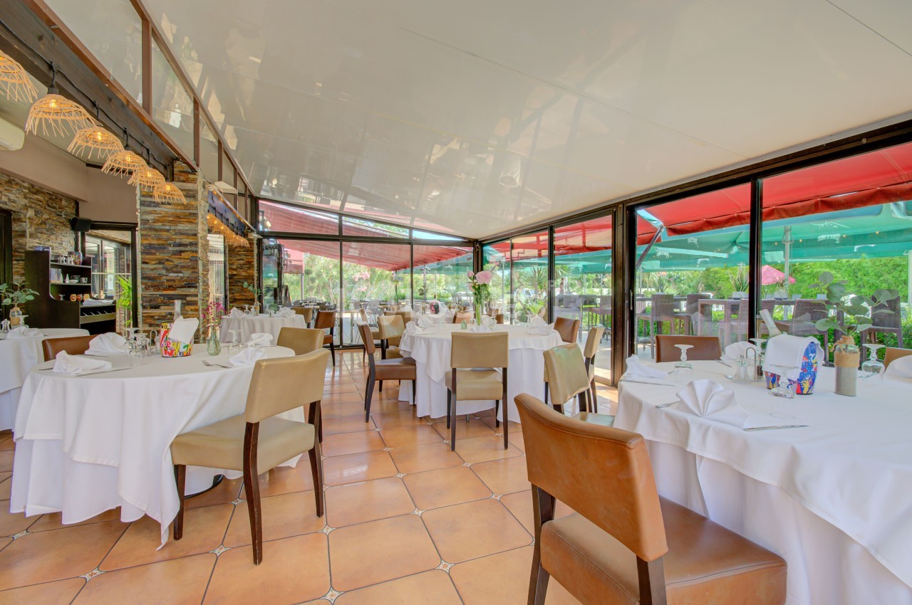 Salle de restaurant lumineuse avec tables dressées, nappes blanches, chaises en bois et grandes baies vitrées donnant sur l…