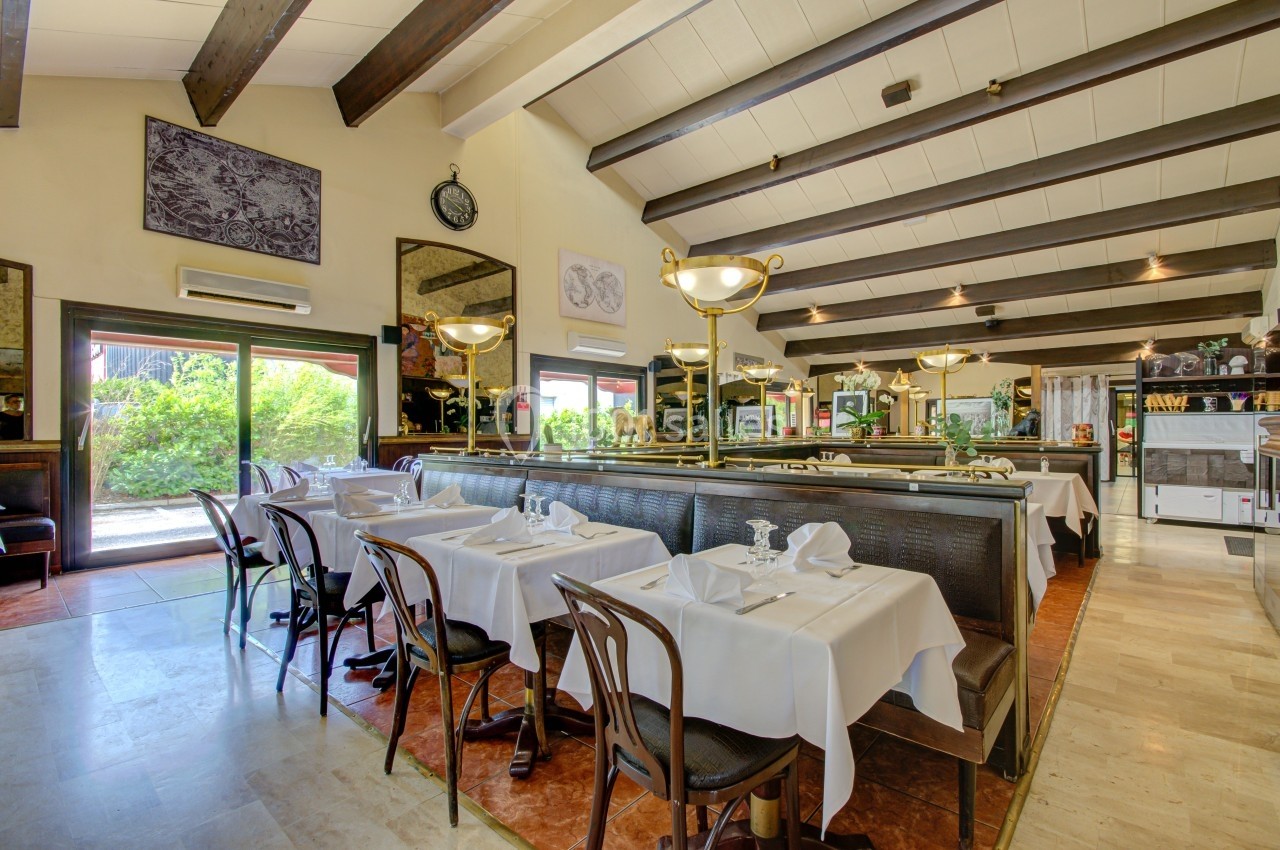 Salle de restaurant lumineuse avec tables dressées, chaises en bois et grandes baies vitrées donnant sur un espace extérieur…