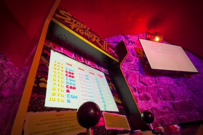 Fléchettes colorées dans une caisse en bois au premier plan, cible de jeu accrochée sur un mur en pierre en arrière-plan.