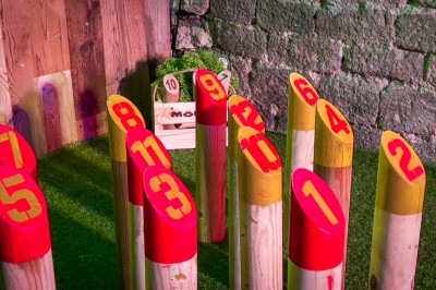 Fléchettes colorées dans une caisse en bois au premier plan, cible de jeu accrochée sur un mur en pierre en arrière-plan.