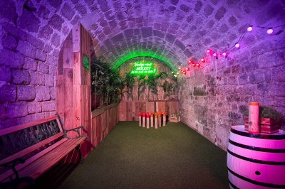 Fléchettes colorées dans une caisse en bois au premier plan, cible de jeu accrochée sur un mur en pierre en arrière-plan.