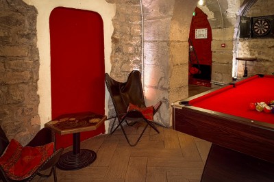 Fléchettes colorées dans une caisse en bois au premier plan, cible de jeu accrochée sur un mur en pierre en arrière-plan.