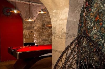 Fléchettes colorées dans une caisse en bois au premier plan, cible de jeu accrochée sur un mur en pierre en arrière-plan.