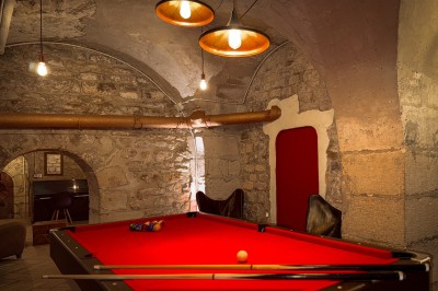 Fléchettes colorées dans une caisse en bois au premier plan, cible de jeu accrochée sur un mur en pierre en arrière-plan.