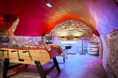 Fléchettes colorées dans une caisse en bois au premier plan, cible de jeu accrochée sur un mur en pierre en arrière-plan.