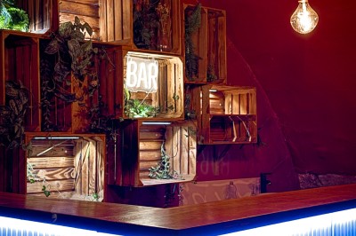 Fléchettes colorées dans une caisse en bois au premier plan, cible de jeu accrochée sur un mur en pierre en arrière-plan.