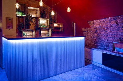 Fléchettes colorées dans une caisse en bois au premier plan, cible de jeu accrochée sur un mur en pierre en arrière-plan.