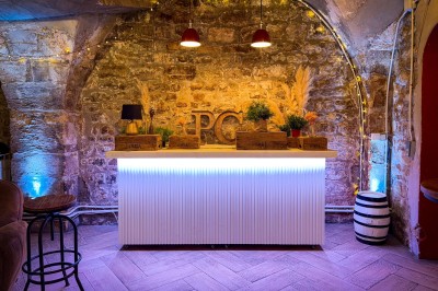 Fléchettes colorées dans une caisse en bois au premier plan, cible de jeu accrochée sur un mur en pierre en arrière-plan.