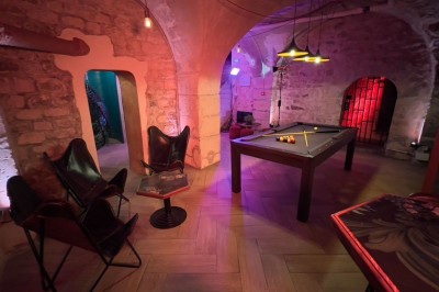 Fléchettes colorées dans une caisse en bois au premier plan, cible de jeu accrochée sur un mur en pierre en arrière-plan.