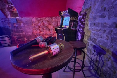 Fléchettes colorées dans une caisse en bois au premier plan, cible de jeu accrochée sur un mur en pierre en arrière-plan.