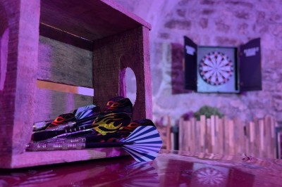 Fléchettes colorées dans une caisse en bois au premier plan, cible de jeu accrochée sur un mur en pierre en arrière-plan.