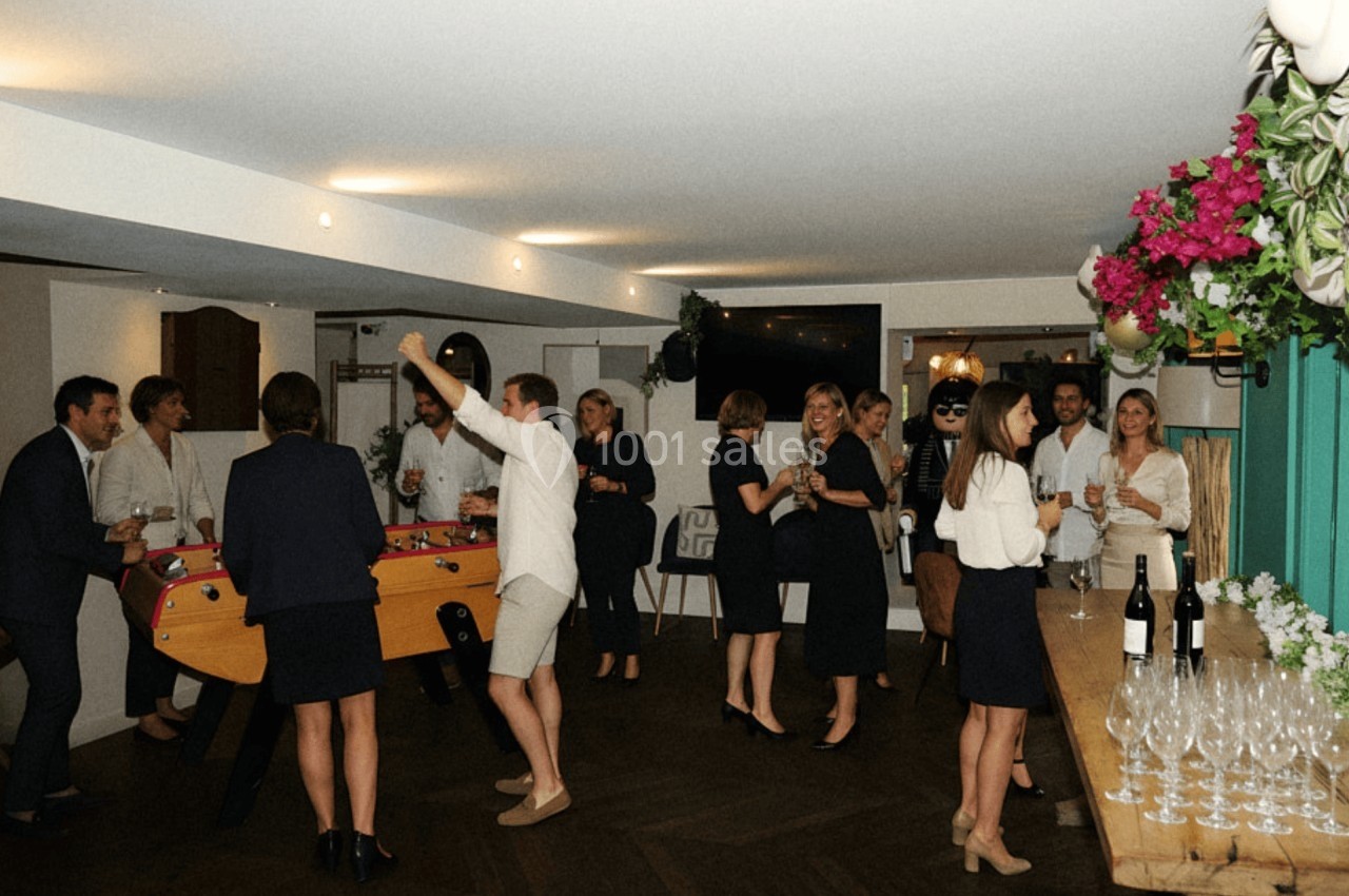 Des personnes discutent et jouent au baby-foot dans une salle lumineuse décorée de fleurs et de tables dressées.
