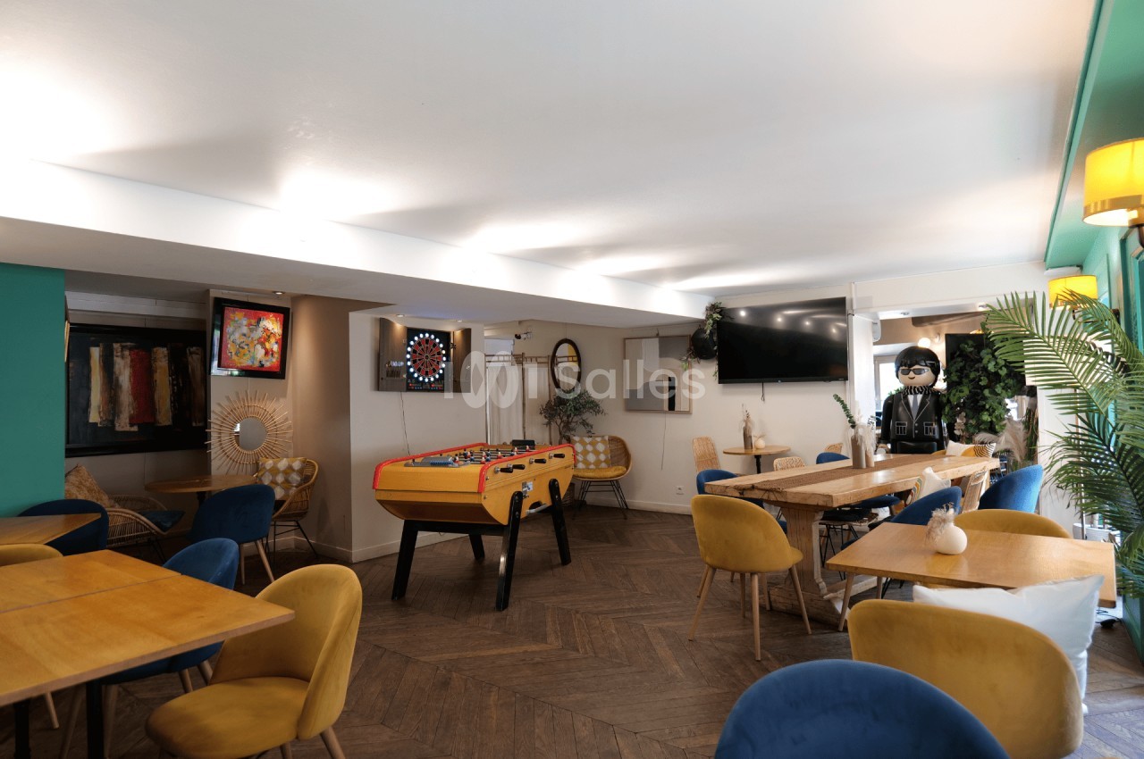 Salle lumineuse avec tables en bois, fauteuils colorés, baby-foot, fléchettes et décoration moderne.