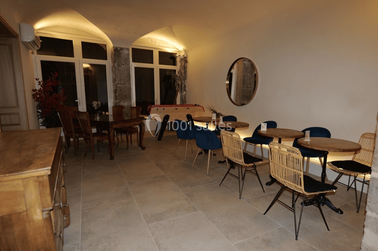 Salle lumineuse avec tables, chaises variées, baby-foot, miroir rond et murs en pierre apparente.