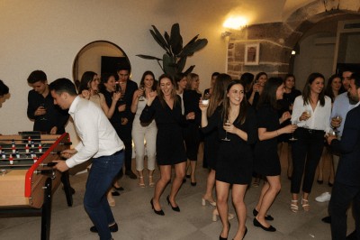 Salle lumineuse avec tables, chaises variées, baby-foot, miroir rond et murs en pierre apparente.
