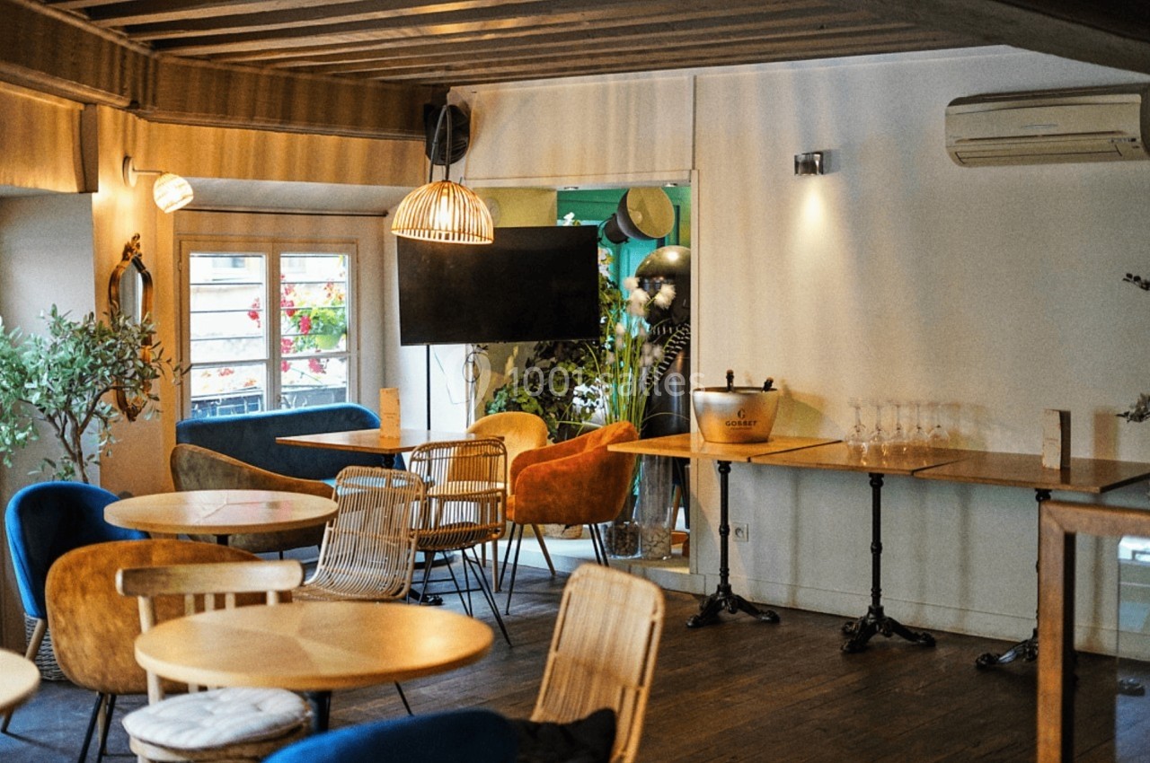 Salle lumineuse avec tables en bois, chaises variées, plantes décoratives et un écran mural.