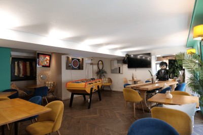 Salle lumineuse avec tables, chaises variées, baby-foot, miroir rond et murs en pierre apparente.