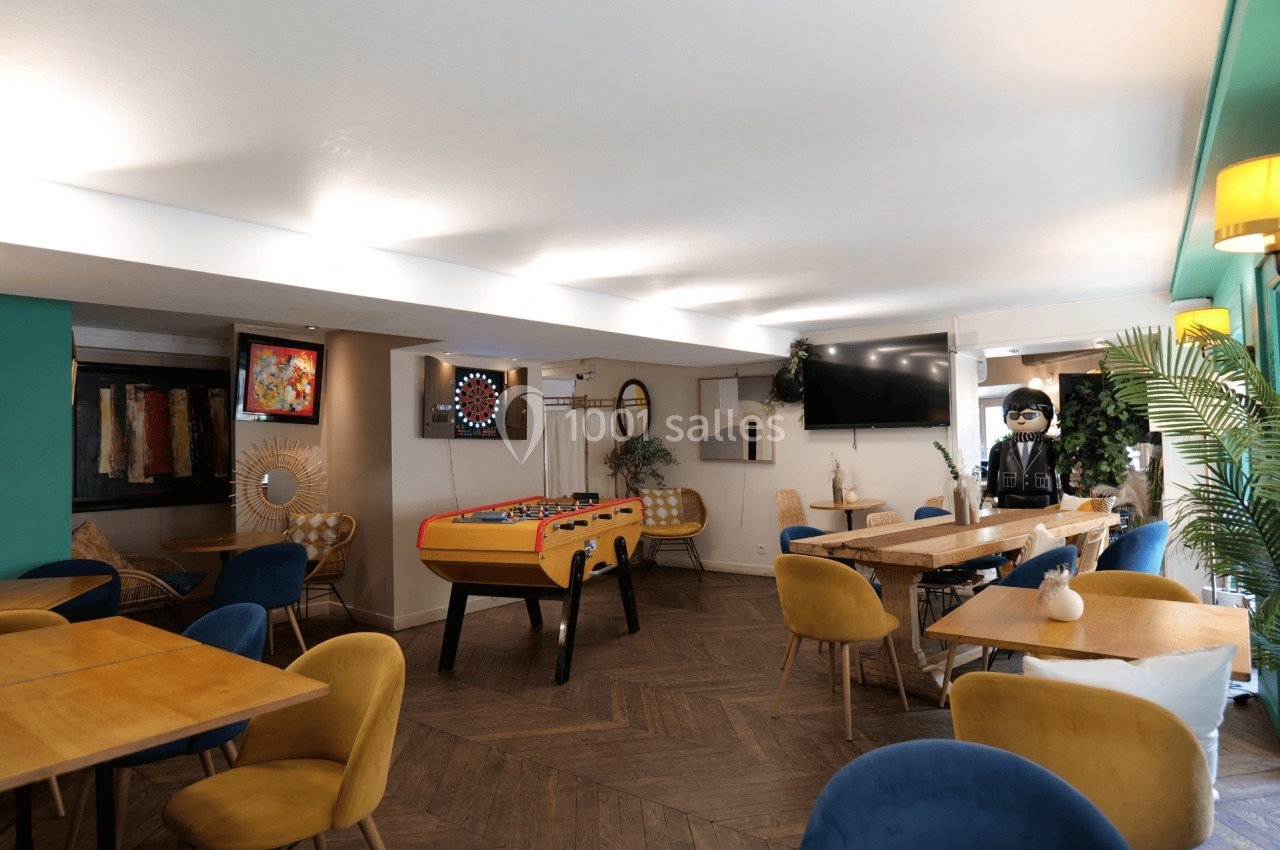 Salle lumineuse avec tables en bois, fauteuils colorés, baby-foot, fléchettes et décoration moderne.