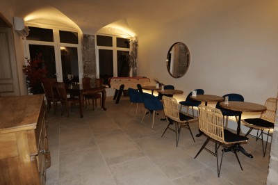 Salle lumineuse avec tables, chaises variées, baby-foot, miroir rond et murs en pierre apparente.