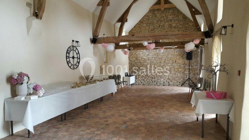 Salle de réception lumineuse avec murs en pierre, poutres apparentes et tables décorées pour un événement.