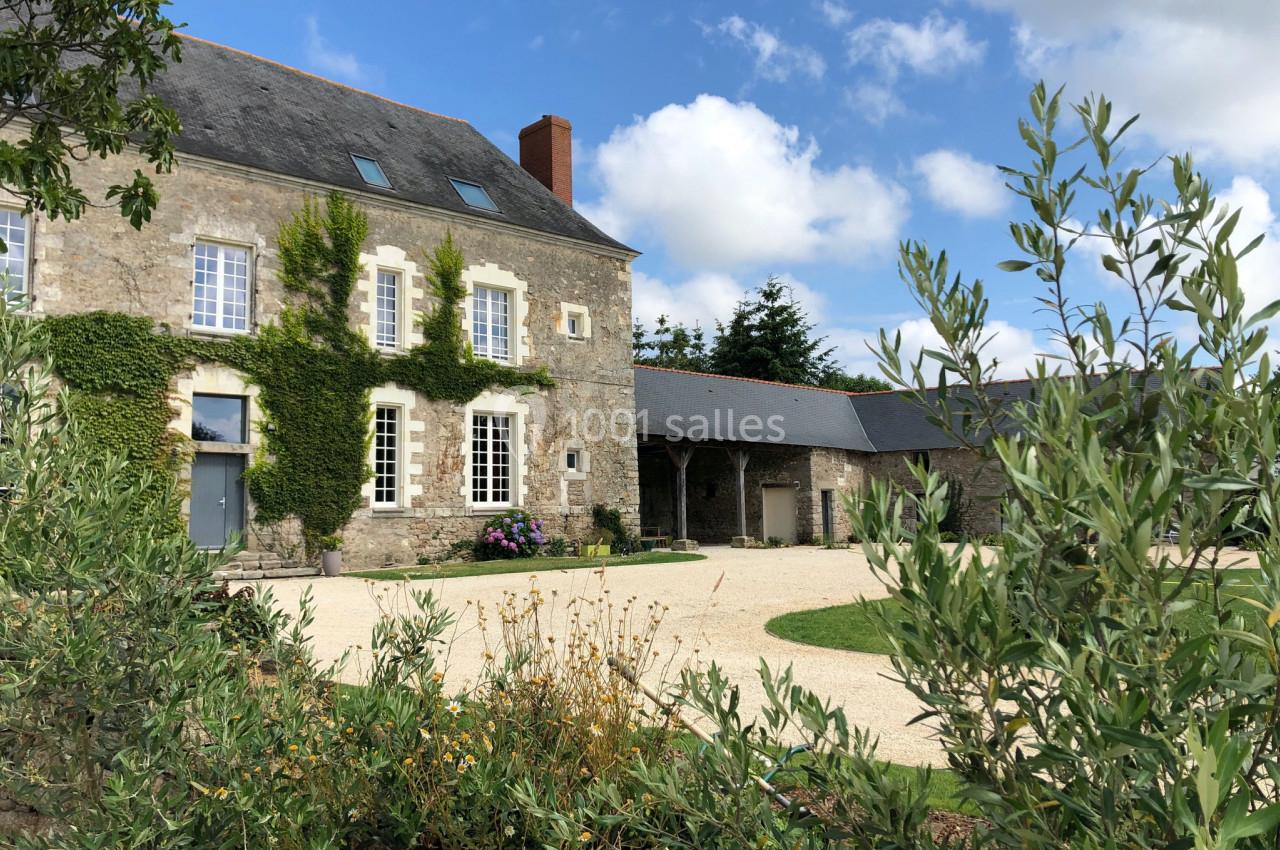 Façade d'une maison en pierre avec des volets blancs, entourée de végétation et d'une cour gravillonnée.