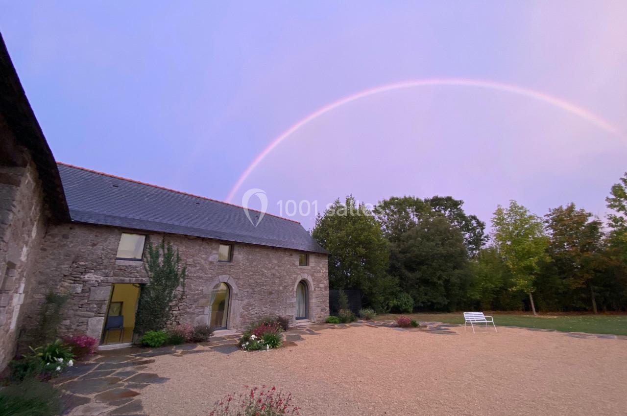 Façade en pierre d'une maison avec un arc-en-ciel dans un ciel pastel, entourée de végétation et d'une cour gravillonnée.