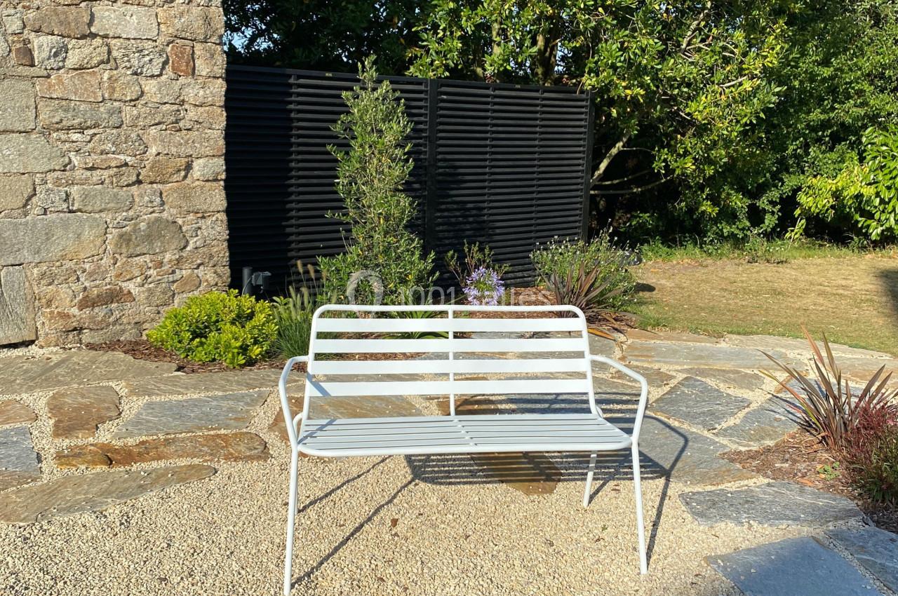 Banc en métal blanc placé sur une terrasse en pierre, entouré de végétation et d'un mur en pierre.
