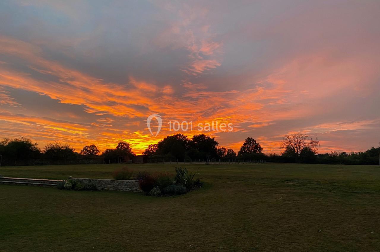 Ciel aux teintes orange et rose au coucher du soleil, avec un grand espace vert et des arbres à l'horizon.